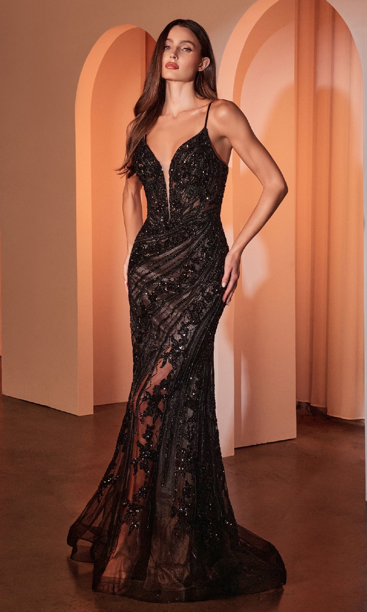 A woman stands indoors wearing the Ladivine CC0389, a long black prom dress featuring beaded lace, sheer panels, and thin straps.